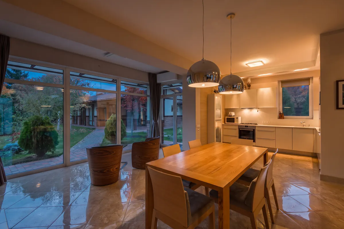 Edición con IA de día a atardecer a las 22:00: cocina abierta con comedor y ventanales de suelo a techo hacia el jardín