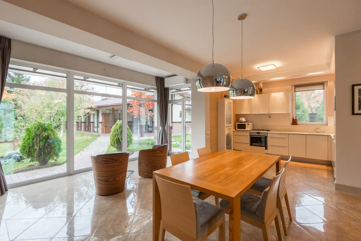Foto diurna original: cocina abierta con comedor y ventanales de suelo a techo hacia el jardín